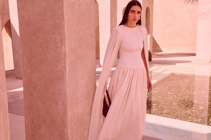 Elegant woman in a long, off-white dress with cape sleeves stands in a sunlit, colonnaded courtyard, exuding sophistication and style.