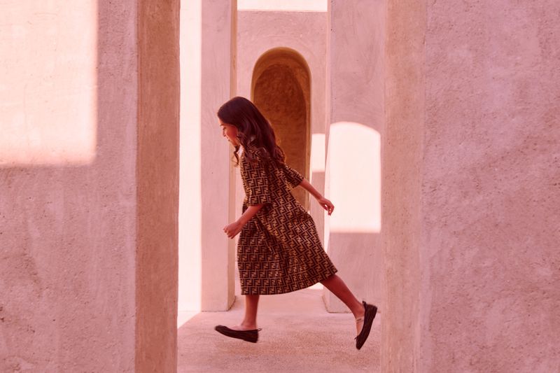 A young girl in a patterned dress strides through a corridor with light pink walls, creating a dynamic, eye-level fashion portrait.