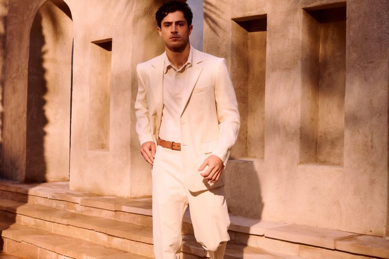 Man wearing a cream suit and brown belt standing outdoors against a stone architectural building exterior with warm sunlight