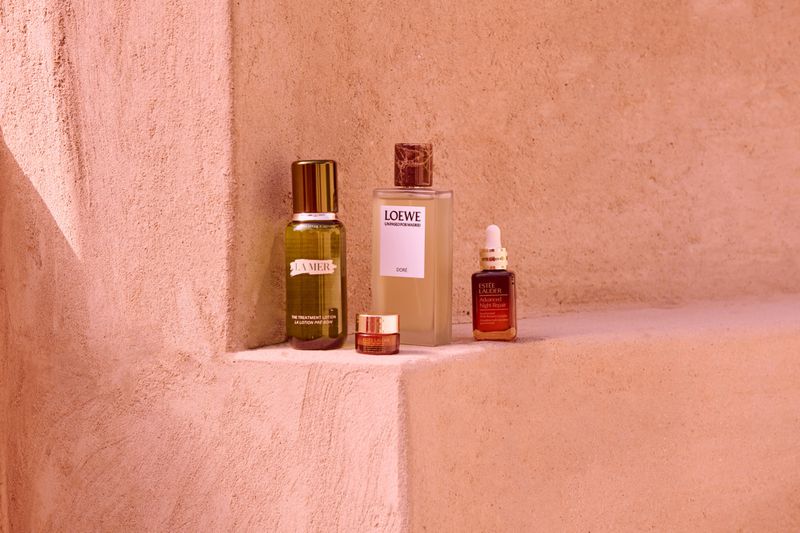 A collection of La Mer, Loewe, and Estee Lauder skincare bottles are arranged on a stucco surface, bathed in soft, warm lighting.