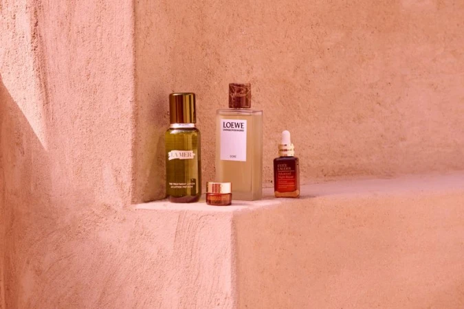 A collection of La Mer, Loewe, and Estee Lauder skincare bottles are arranged on a stucco surface, bathed in soft, warm lighting.