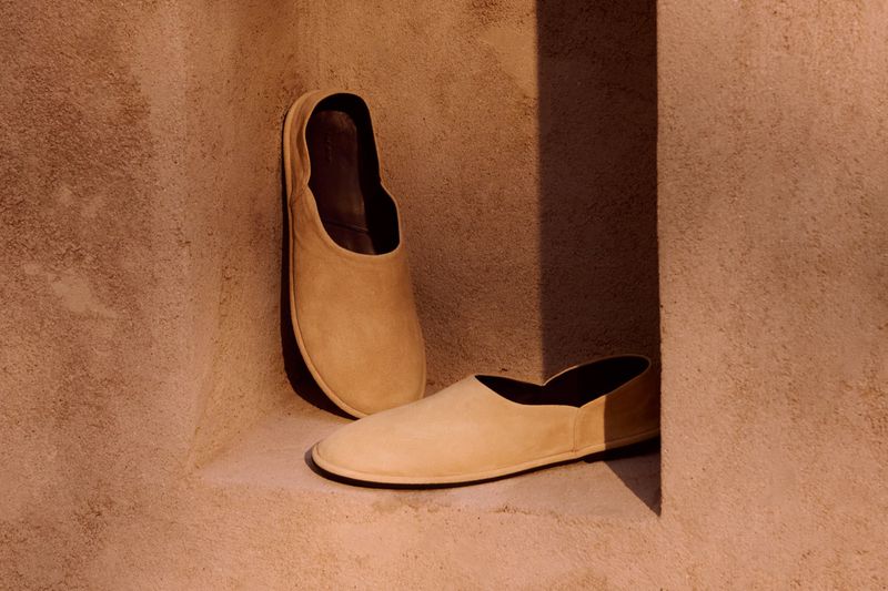 Minimalist tan-colored leather slip-on shoes staged in a shallow stone alcove with deep shadows; the shoes exhibit fine craftsmanship.