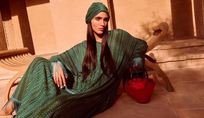 Fashion shot of a woman wearing a green patterned dress and head wrap, seated next to a red purse against a tan wall backdrop.