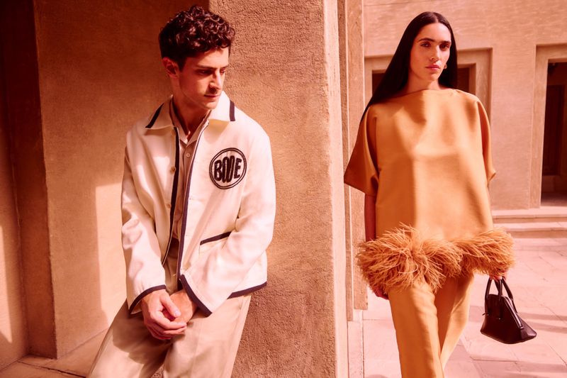 Fashion shot of a stylish couple in a building exterior; the male wears a cream-colored jacket while the female is in brown top and pants.
