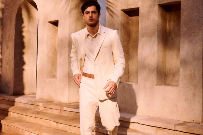 Man wearing a cream suit and brown belt standing outdoors against a stone architectural building exterior with warm sunlight