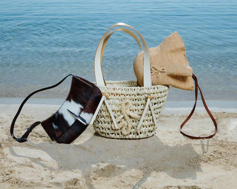 Three fashion handbags on a sandy beach with a blue sea background. The bags include a straw tote and brown and white leather bag.