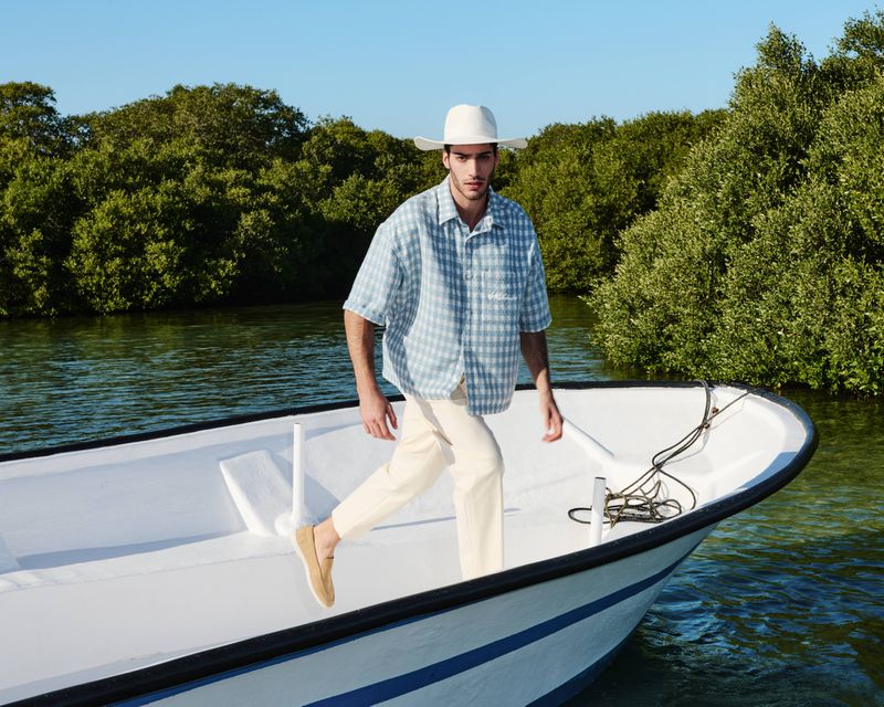 A man in a hat walks along the bow of a white boat on the water, trees visible in the background on a bright sunny day.