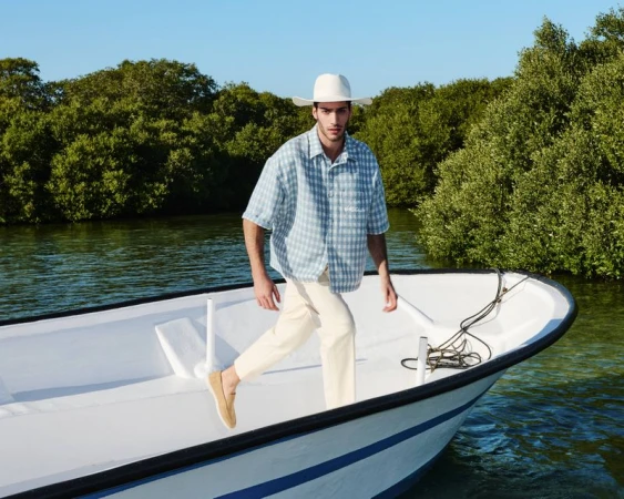 A man in a hat walks along the bow of a white boat on the water, trees visible in the background on a bright sunny day.
