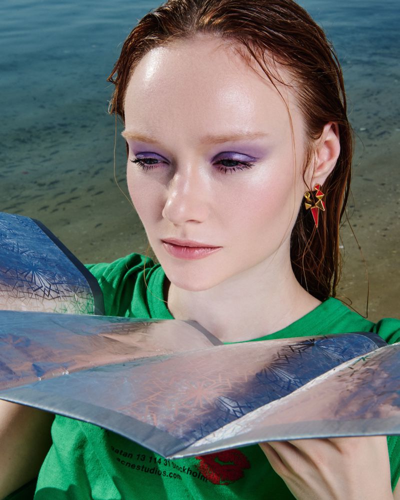 Portrait of a red-haired woman with purple eyeshadow, wearing a green t-shirt and earrings, posing with reflective material, beach background.