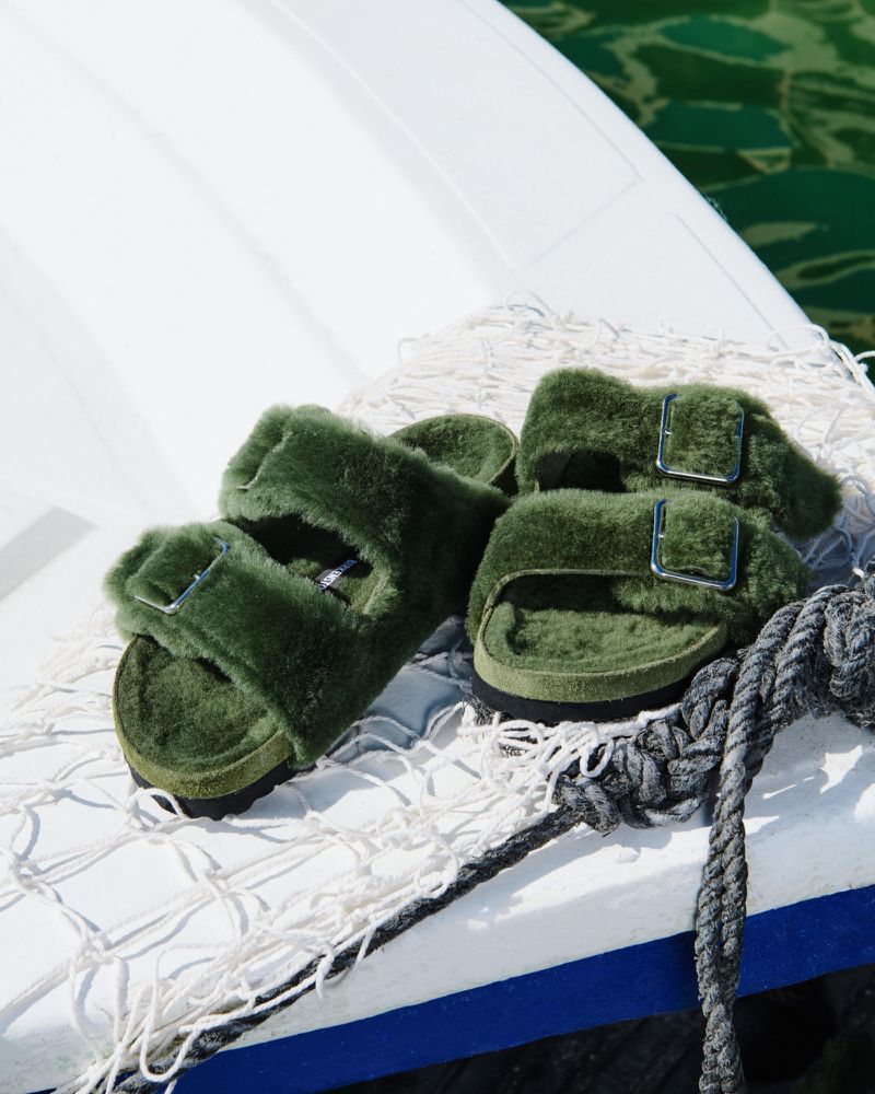 Olive green, shearling slide sandals with silver buckles sit atop a white boat surface, with a white net and a gray rope.