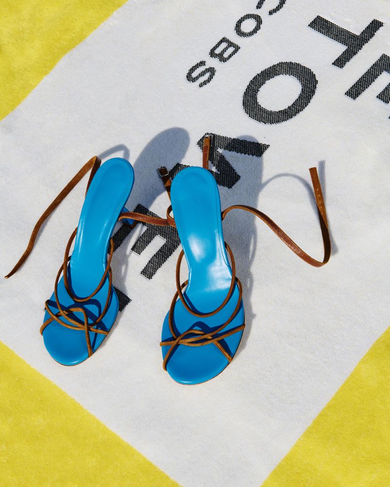 Eye-level view of blue high-heel sandals with brown straps on a diagonally-striped yellow and white towel with black brand text.