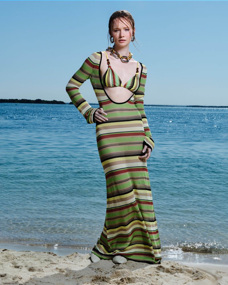 A fashionable adult woman stands on a sandy beach, wearing a striped knit dress, posing in front of ocean water under a clear sky.