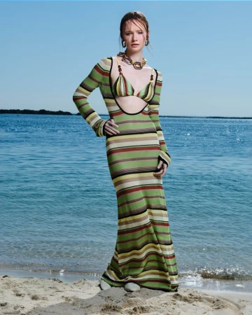 A fashionable adult woman stands on a sandy beach, wearing a striped knit dress, posing in front of ocean water under a clear sky.