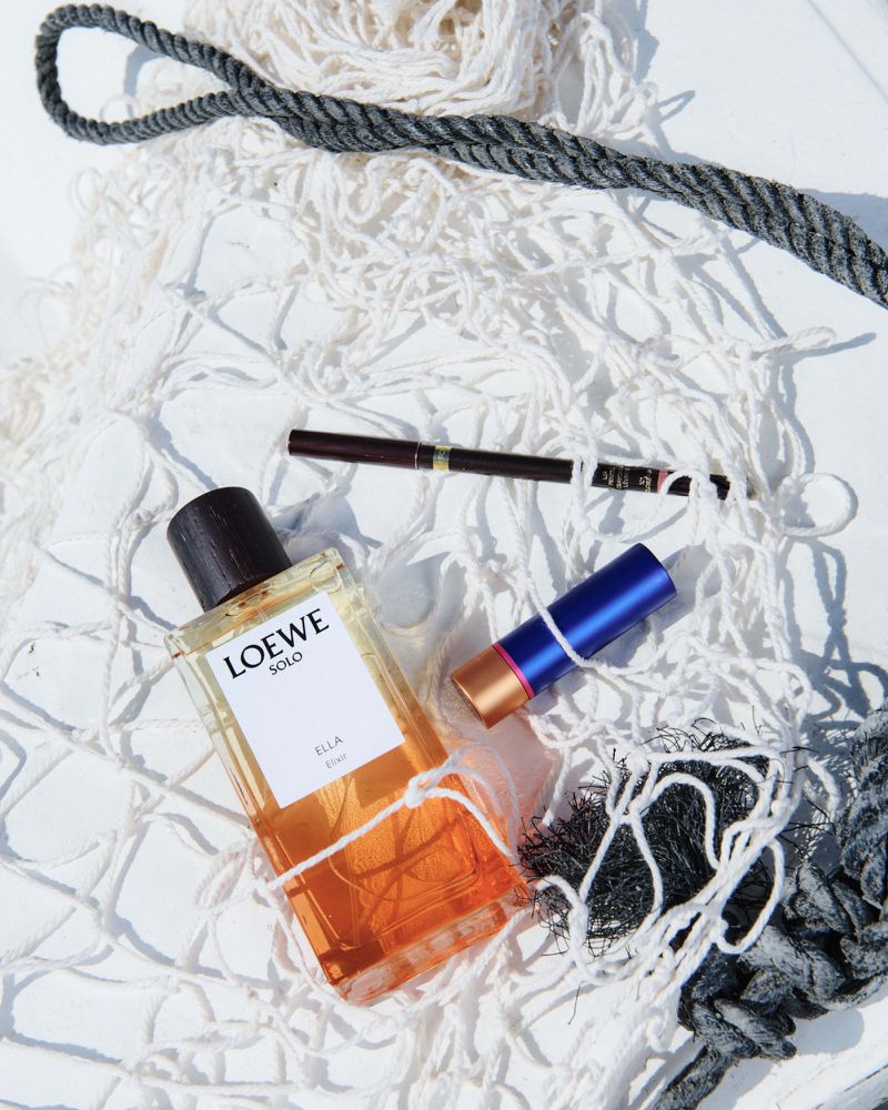 Still life flat lay of a Loewe Solo Ella perfume bottle, an eyeliner, and lipstick against a white netting with a grey rope accent.