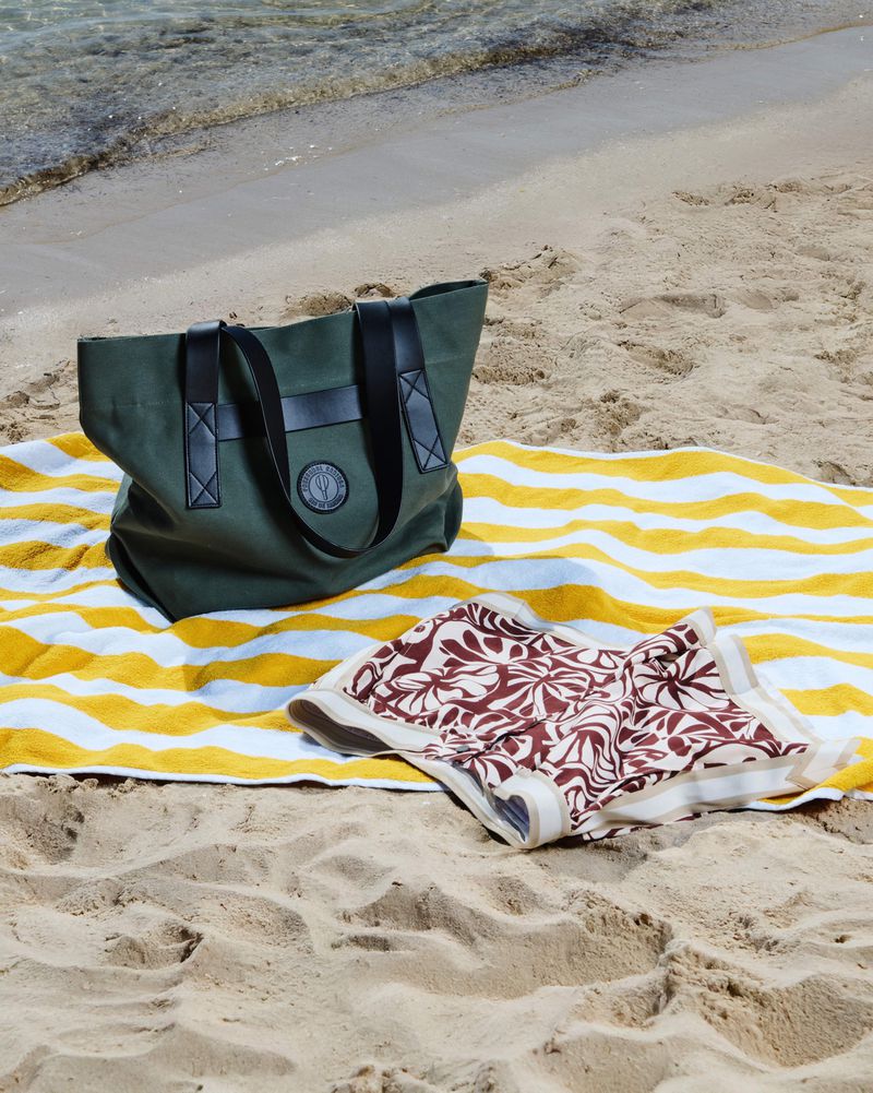 Green tote bag with black straps and round logo on a striped yellow and white beach towel with a patterned sarong, outdoor beach setting.