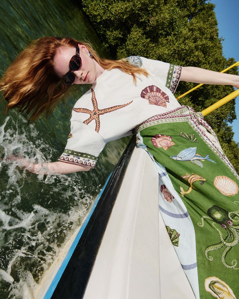 Red-haired woman in sea-themed top on a boat, wearing sunglasses. She is reaching out towards the water in bright daylight.