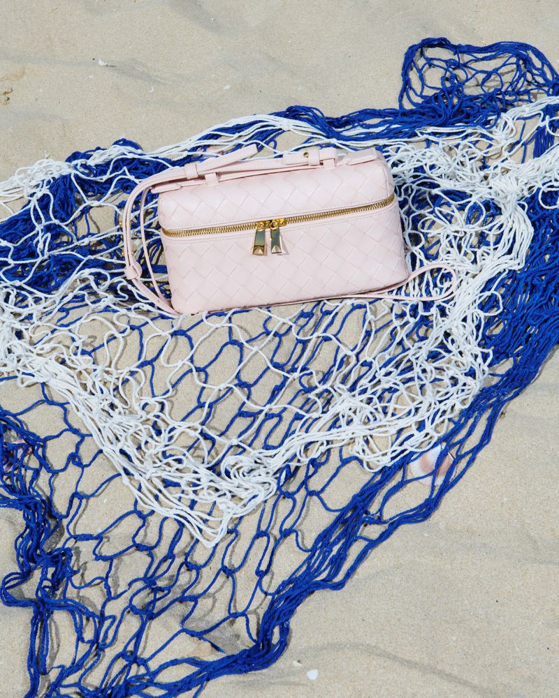 Pink woven handbag with gold zippers on a blue and white net against a tan sand background, shot outdoors for fashion and style.