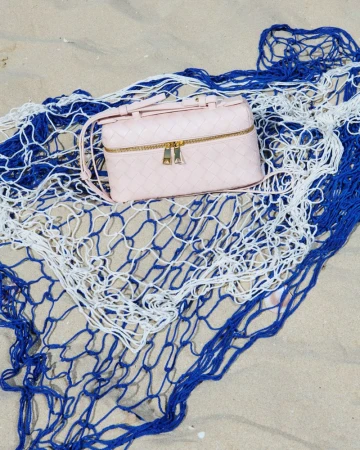 Pink woven handbag with gold zippers on a blue and white net against a tan sand background, shot outdoors for fashion and style.