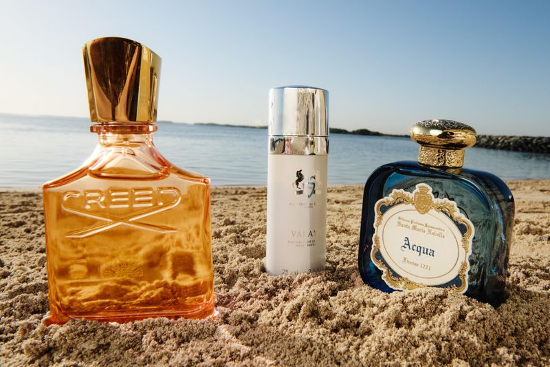 Three perfume bottles on a sandy beach with the ocean and sky in the background under bright daylight, promoting fragrance. 