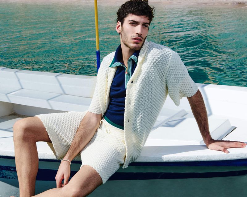A fashionable adult male model poses on a boat in the ocean wearing a cream-colored knit shirt and shorts, set against a backdrop of turquoise water.