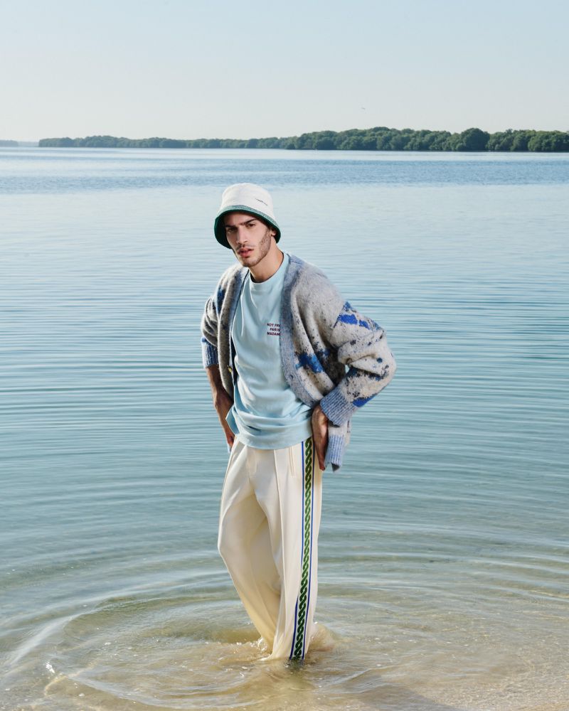 A young adult male model stands in shallow water at the shore, wearing a pastel blue tee, light cardigan, white pants, and a bucket hat.
