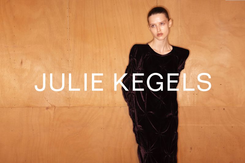 A slender woman poses against a wood paneled wall, modeling a long, dark velvet dress with shoulder pads and gathered seams.
