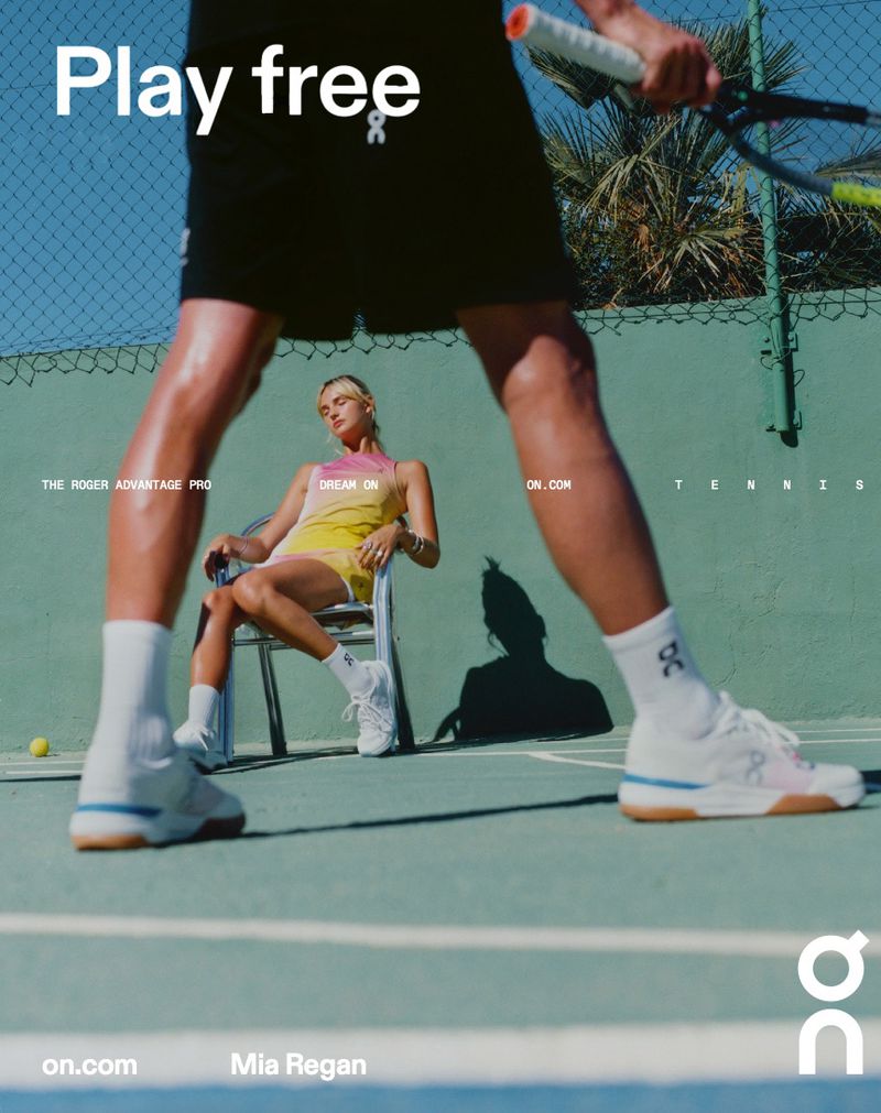 Two tennis players, one standing with a racket and one seated on a chair, on a blue and green outdoor tennis court on a sunny day.
