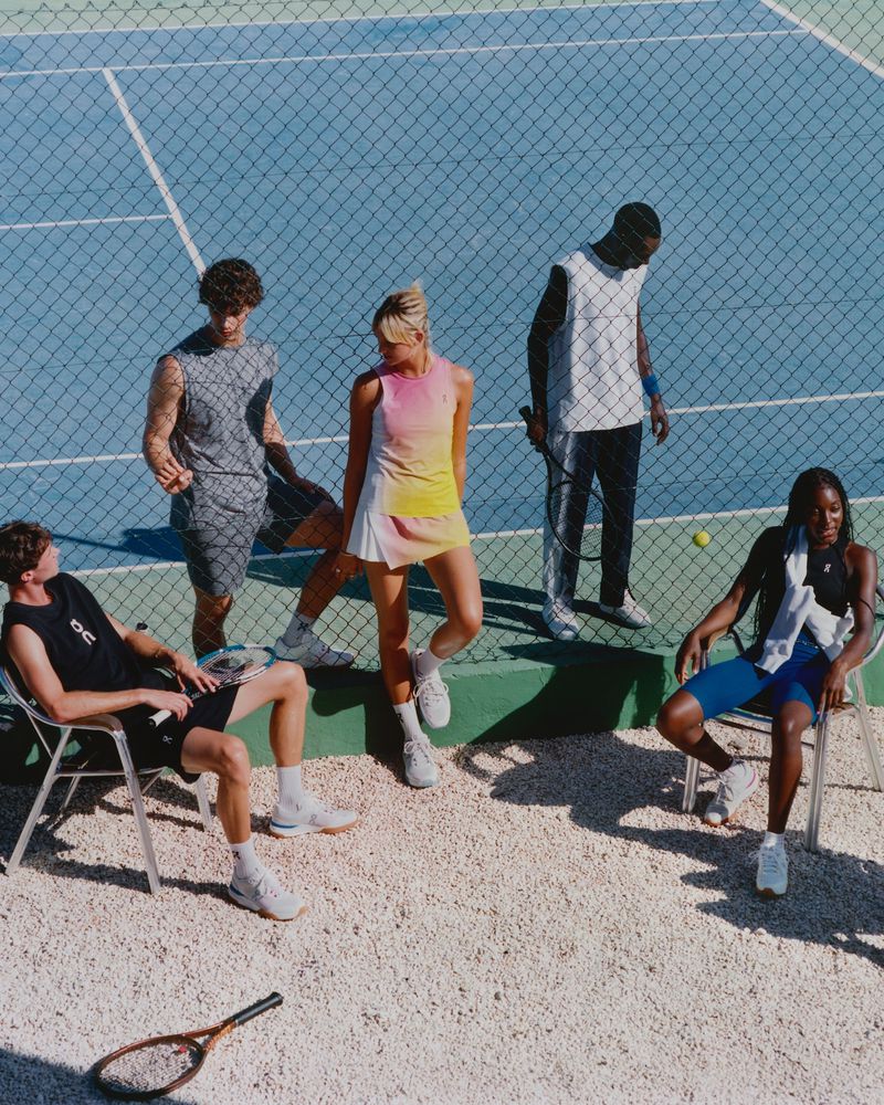Group of diverse athletes gather on a bright blue tennis court. Some sit on chairs near a net while others stand. Fashion and athletic wear are visible.