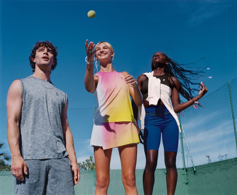 Three athletic people pose on a tennis court under a blue sky. A woman tosses a tennis ball. The models wear sportswear apparel.