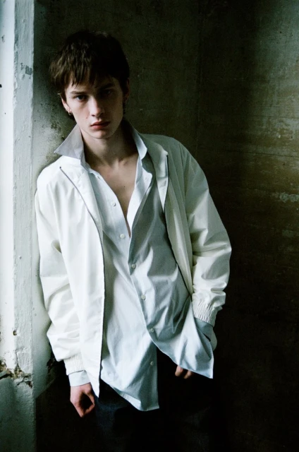 Portrait of a young adult man in an unbuttoned white shirt and a light jacket, leaning against a weathered, grey wall in a shadowy indoor corner.