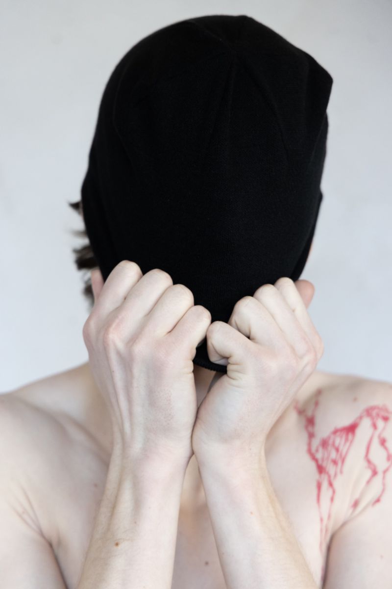 A person wearing a black beanie covers their face with their hands, with a red abstract design visible on their shoulder against a plain background.