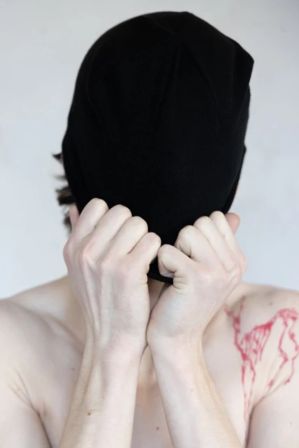 A person wearing a black beanie covers their face with their hands, with a red abstract design visible on their shoulder against a plain background.