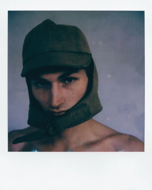 Portrait of a young adult male in a dark green cap with ear flaps, against a light gray backdrop, captured in a Polaroid-style frame.
