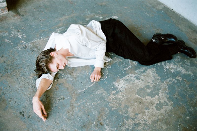Low angle view of a stylish man lying on a concrete floor in a bright white shirt and black pants with buckled shoes.