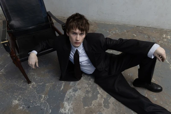 Model wearing a black suit and tie sitting on a weathered floor next to a dark leather chair