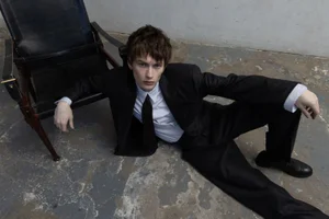 Model wearing a black suit and tie sitting on a weathered floor next to a dark leather chair