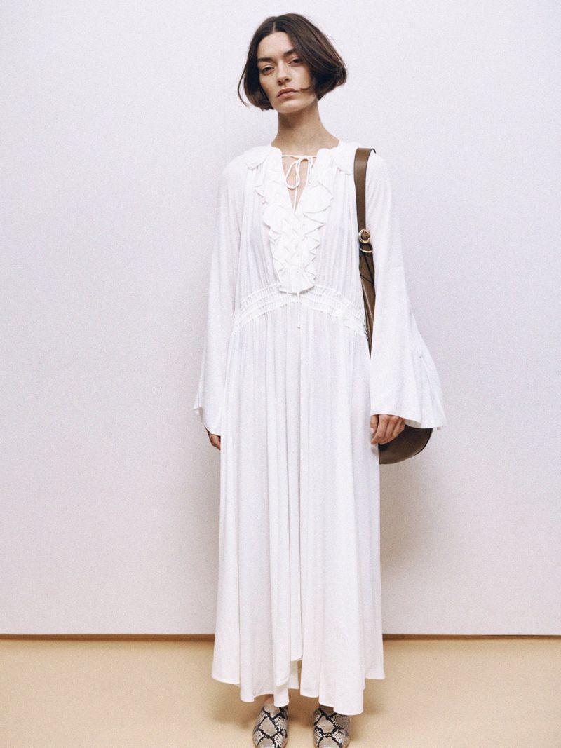 Brunette woman in a white, ruffled, long-sleeve dress with a brown purse and snakeskin shoes, posing against a white background.