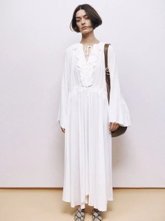 Brunette woman in a white, ruffled, long-sleeve dress with a brown purse and snakeskin shoes, posing against a white background.