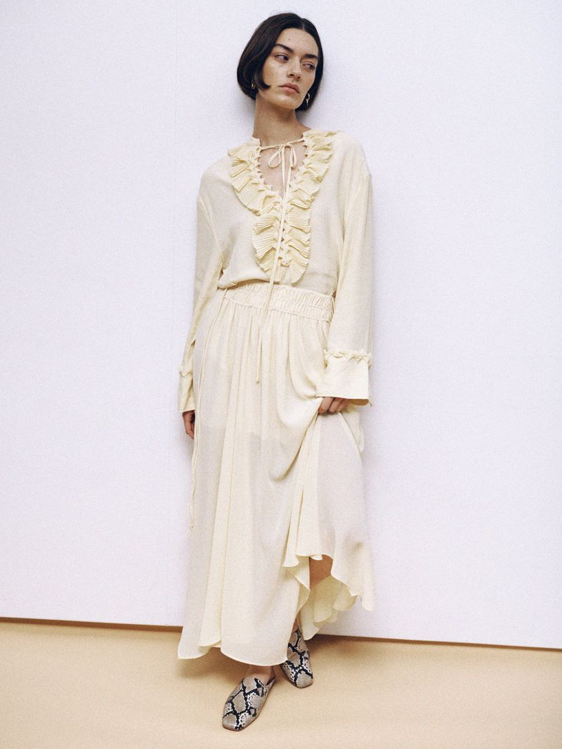A woman in a cream-colored long-sleeved dress and snake-print shoes stands in front of a neutral background, showcasing a fashion style.