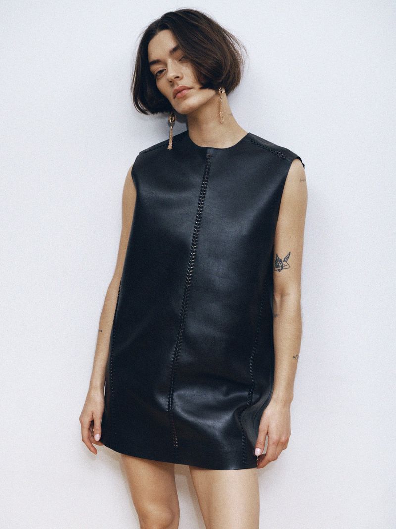 A woman with short dark hair poses in a sleeveless black leather mini dress against a plain white background, showcasing a minimalist fashion style.