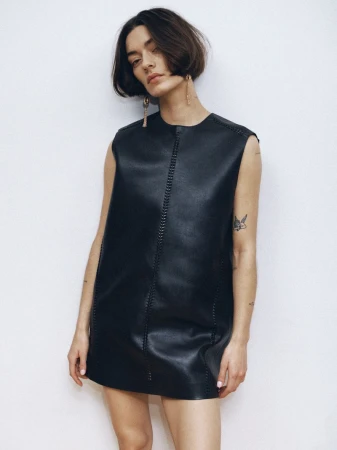 A woman with short dark hair poses in a sleeveless black leather mini dress against a plain white background, showcasing a minimalist fashion style.