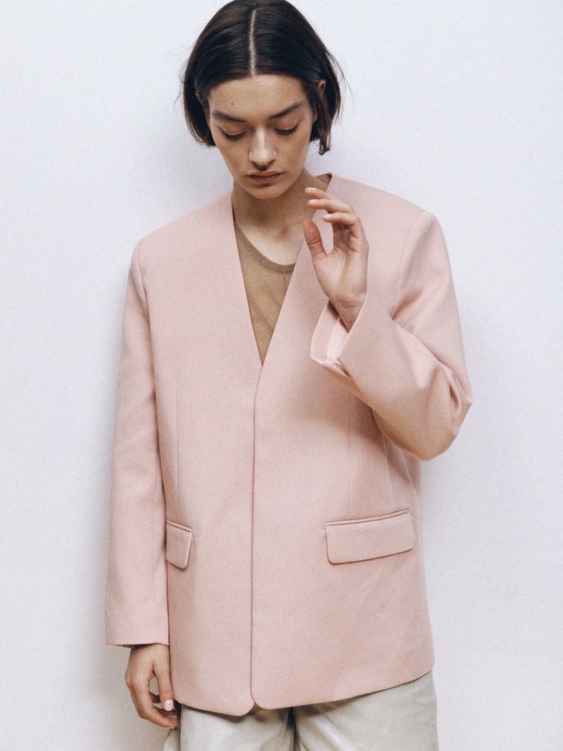 Woman in a light pink blazer and neutral trousers poses against a white wall in a fashion editorial style, looking downward.