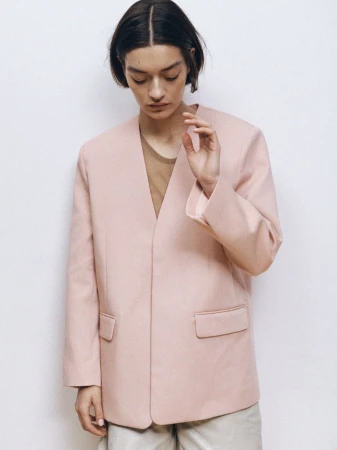 Woman in a light pink blazer and neutral trousers poses against a white wall in a fashion editorial style, looking downward.