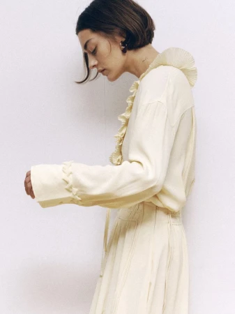 Fashion shot of a woman in a cream-colored blouse with ruffle collar and a skirt, standing in front of a white wall, side view.