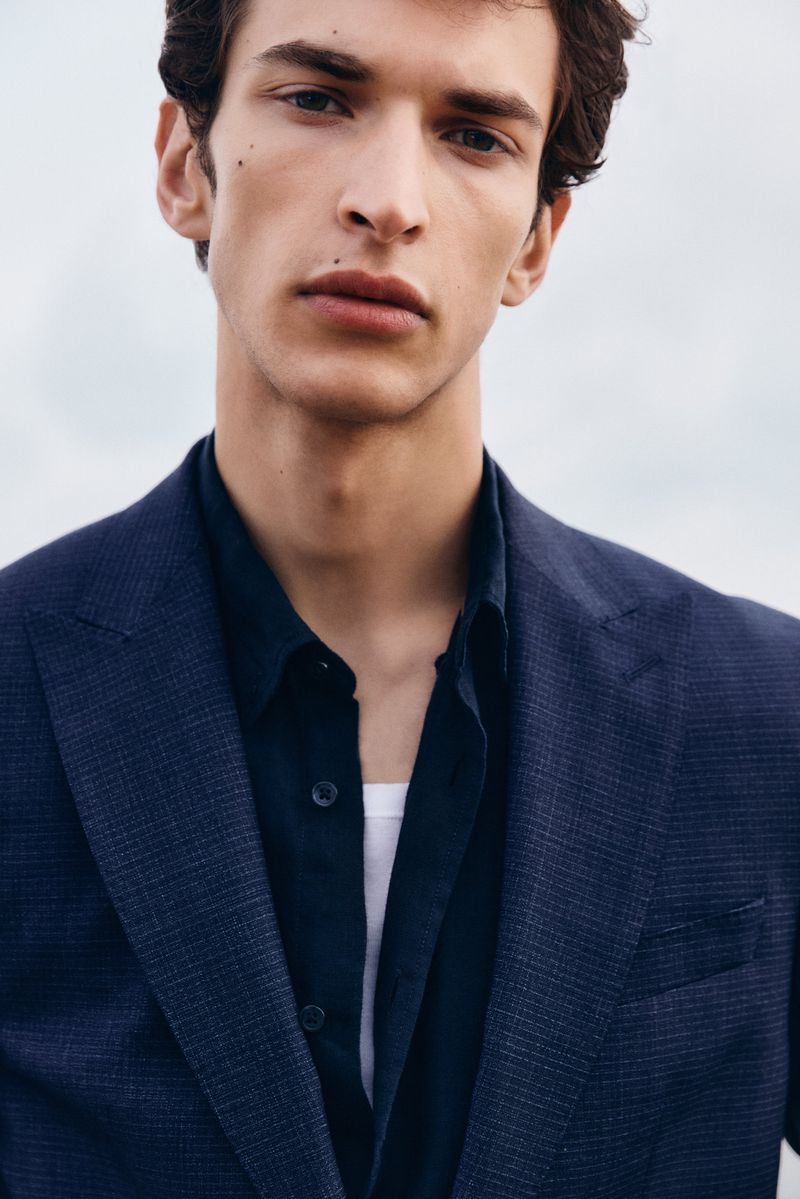 Close-up portrait of a male model wearing a navy blazer and dark button-down shirt against an overcast sky.