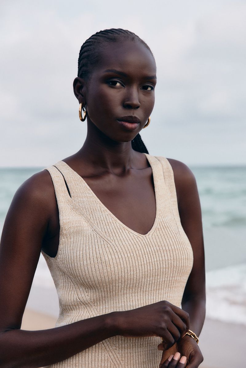 Close-up of a Black woman with braided hair wearing a beige dress and gold earrings posing on a beach with the ocean in the background.