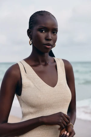 Close-up of a Black woman with braided hair wearing a beige dress and gold earrings posing on a beach with the ocean in the background.