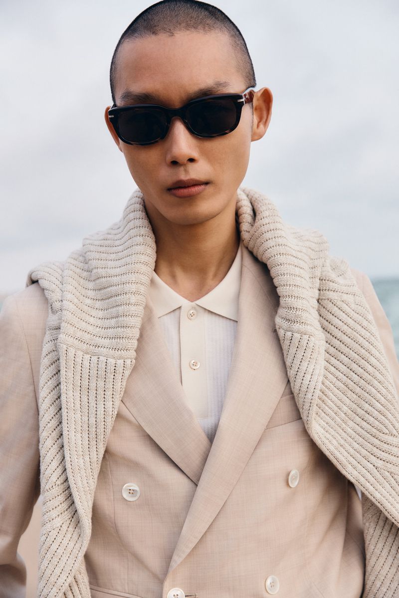 Model wearing dark sunglasses, beige double-breasted suit jacket, and cream sweater draped over shoulders outdoors