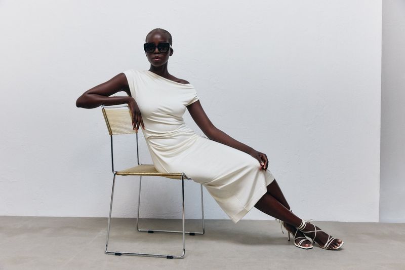 Model Ajok Madel wearing a white dress seated on a metal frame chair against a plain white studio backdrop.