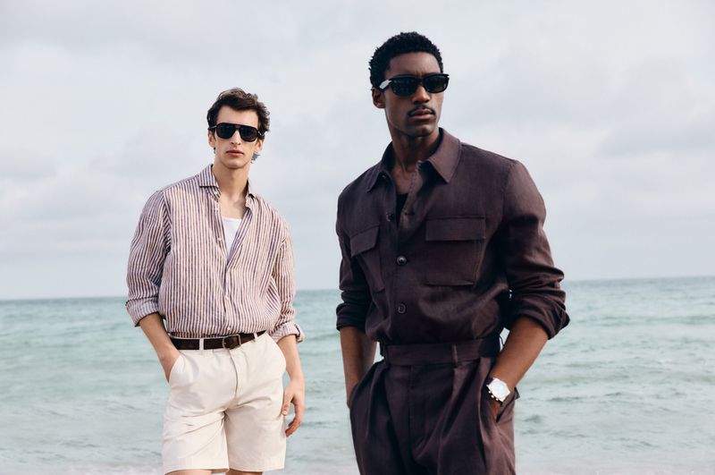 Two adult men pose on a beach wearing casual summer attire, including button-down shirts, shorts, trousers, and sunglasses.